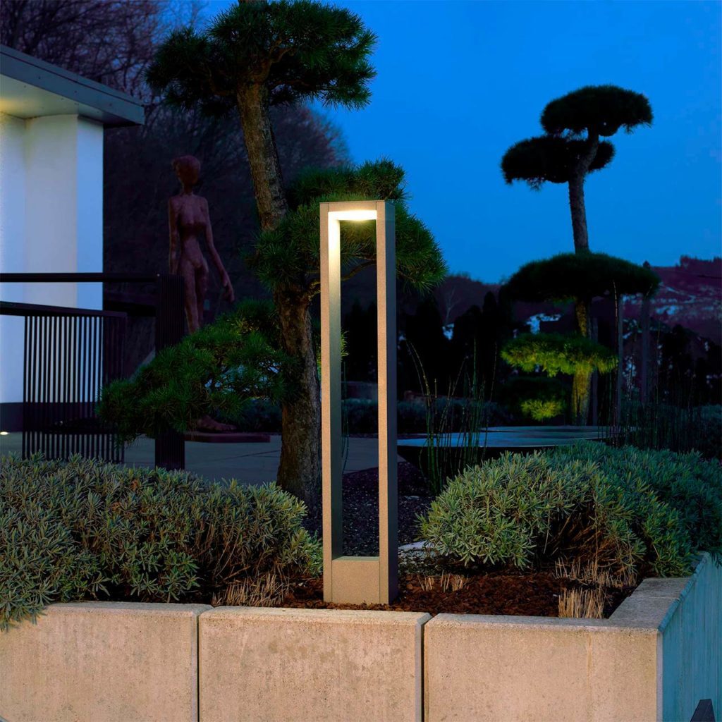 A modern garden light stands at the edge of a flower bed.