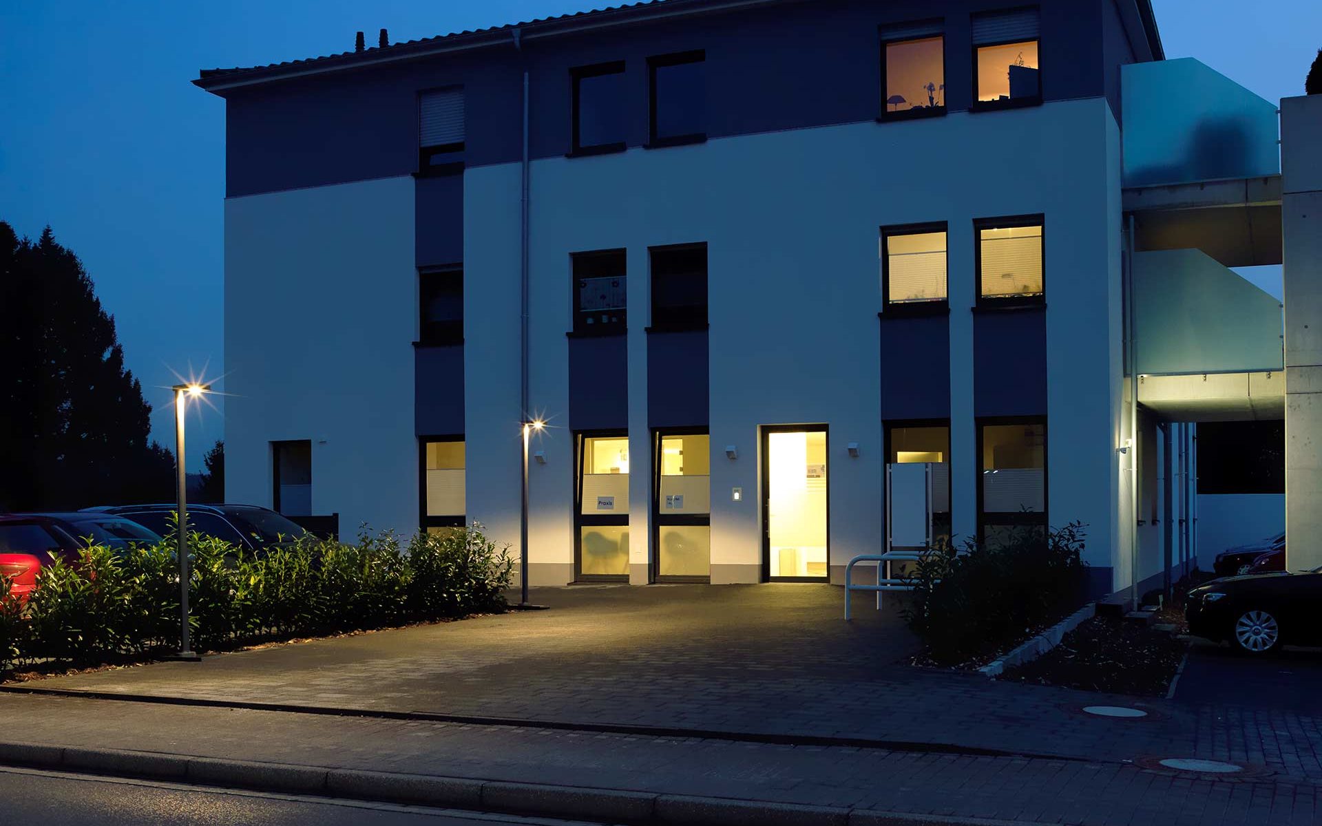 The sidewalk leading to a modern house is lit in the evening by several pole lights that shine all the way to a nearby parking space.