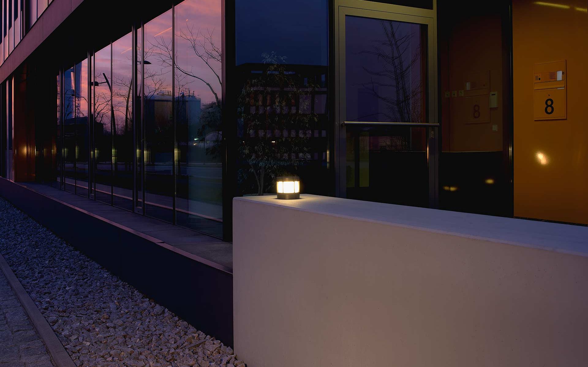 A small pedestal light provides picturesque light on a wall in front of a building complex.